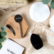 A collection of eco-friendly accessories are arranged on a light brown surface with natural elements. Included are reusable bamboo cotton pads, a black blending sponge and a silver tube squeezer